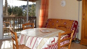 A cozy room with a wooden table and chairs, a sofa with a patterned cover, a large window with a view of trees, and a decorative wall piece. - Cozy 2-Room Apartment for 6 with Terrace, Near Ski Slopes, Pet-Friendly (Bolquère-Pyrénées 2000)