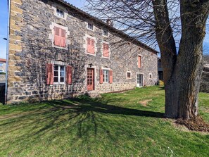 Un edificio de piedra con contraventanas rojas, un patio cubierto de césped y un árbol.