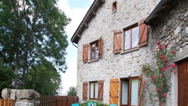 A stone building with wooden shutters, a fenced garden, and a table with chairs.