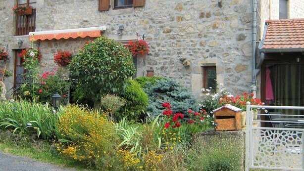 A stone building with a rustic facade, wooden shutters, and a small garden with various plants and flowers.