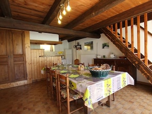 Un comedor rústico con una mesa de madera dispuesta para cuatro personas, rodeada de sillas de madera. Hay una cesta de pan sobre la mesa y una escalera es visible al fondo.
