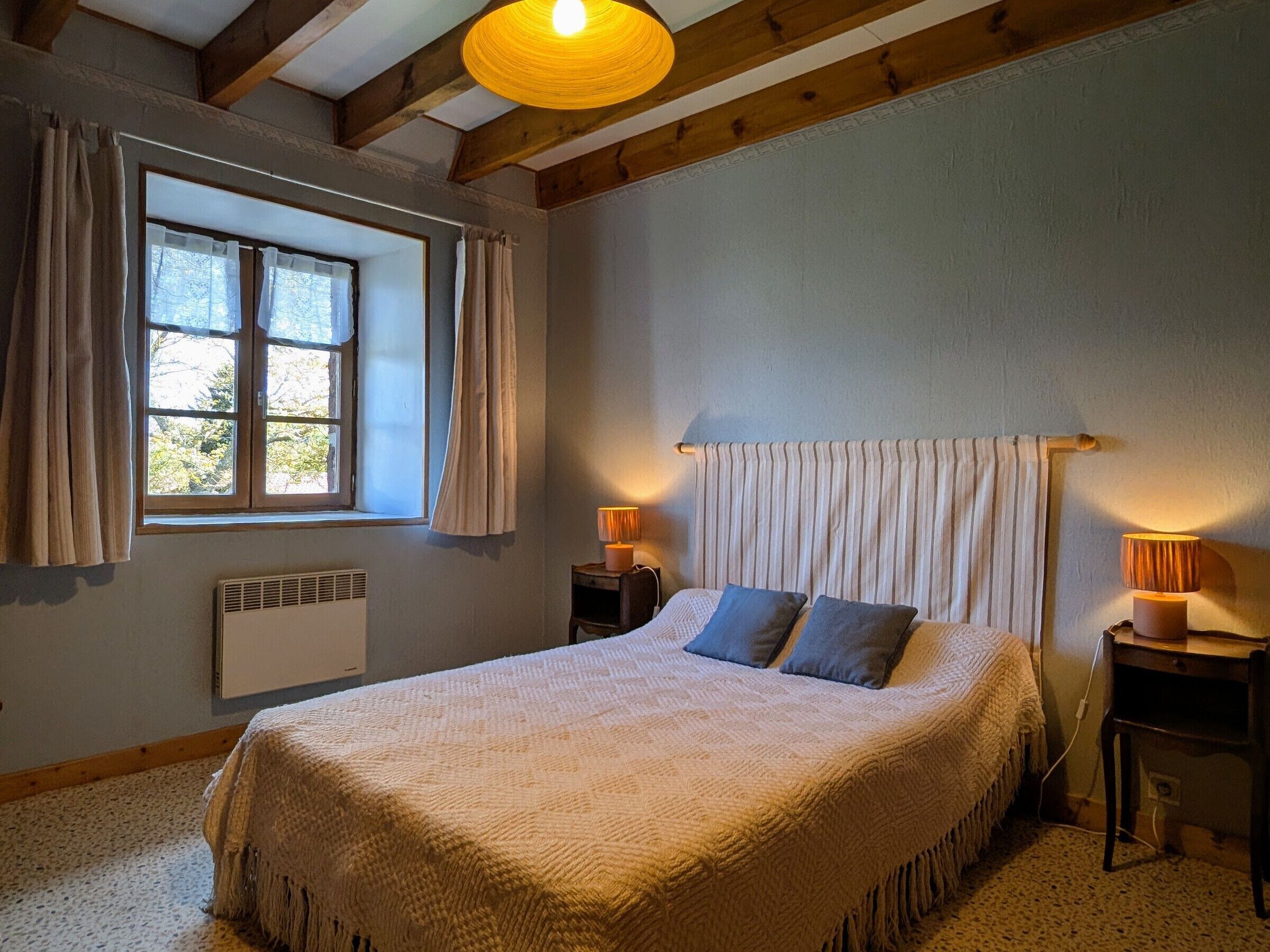 Une chambre à coucher avec un lit, deux lampes de chevet, une fenêtre avec des rideaux, un radiateur et un plafond en bois.