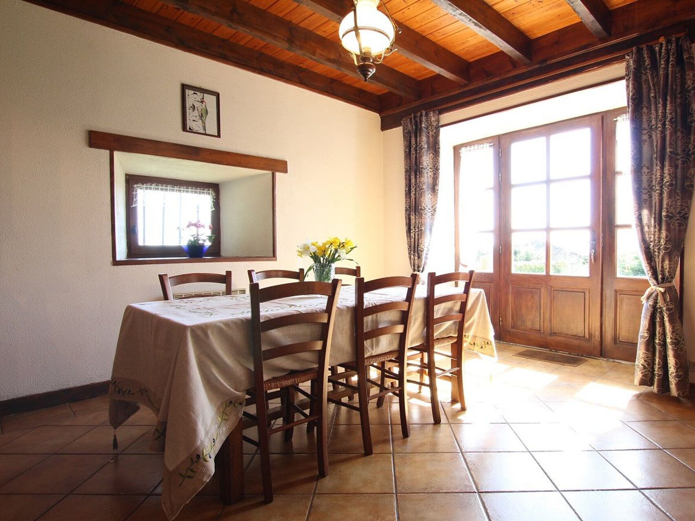 A dining area with a wooden table, chairs, a vase with flowers, a mirror, and a window with curtains.