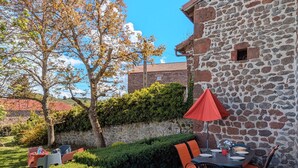 Un bâtiment en pierre doté d’un espace repas comprenant une table et des chaises, un parasol rouge, et offrant une vue sur un arbre et un jardin.