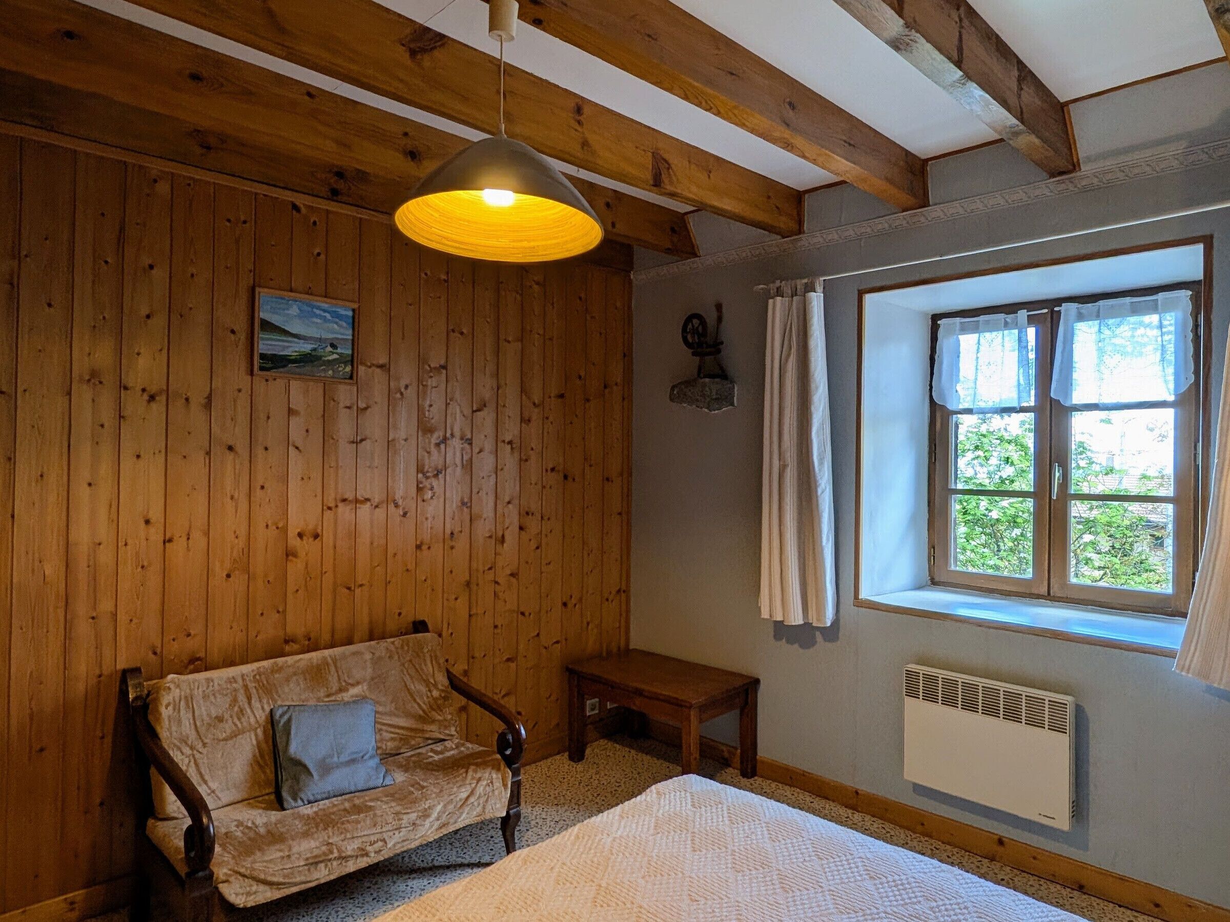 A room with wooden walls, a bed, a bench, a window with curtains, and a hanging light fixture.