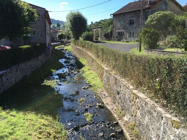 A narrow stone-lined water channel with a grassy bank, flanked by stone buildings and a paved road.