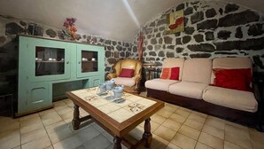 Un salon chaleureux avec un mur en pierre, une armoire verte, une table basse en bois, un canapé beige et un luminaire suspendu.