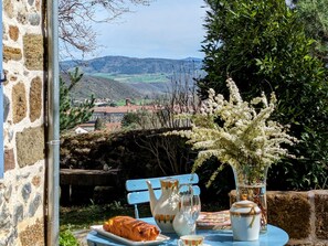 A table with a croissant, a pitcher, a glass, and a vase with white flowers, set against a stone wall with a view of a village and hills. - Vineyard Cabin in Auvergne for Romantic Getaway (Langeac)