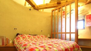 Quarto com estrutura de cama de madeira, colcha xadrez, janela com persianas e prateleira de madeira com livros.