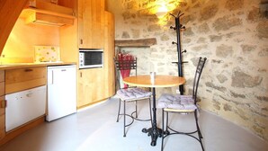 Cozinha acolhedora com armários de madeira, uma geladeira branca e uma parede de pedra. Há uma mesa redonda com duas cadeiras e um abajur.