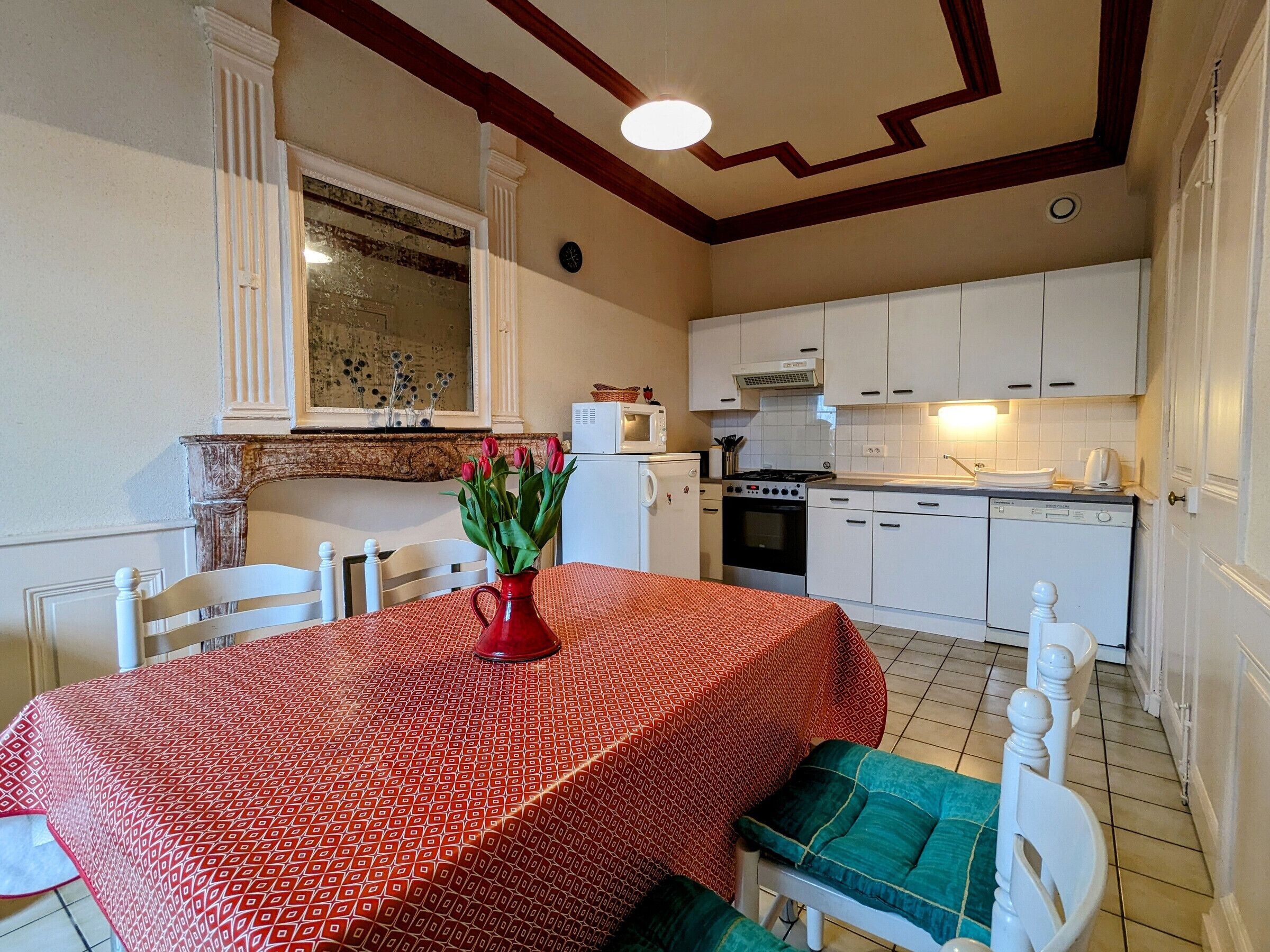 A compact kitchen with white cabinets, a microwave, stove, and a sink. There is a dining table with a red patterned tablecloth and a vase of flowers.