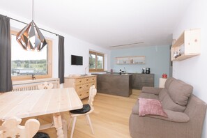 A modern kitchen with wooden cabinets, a dining table, and chairs. There is a sofa with a cushion and a television on the wall.