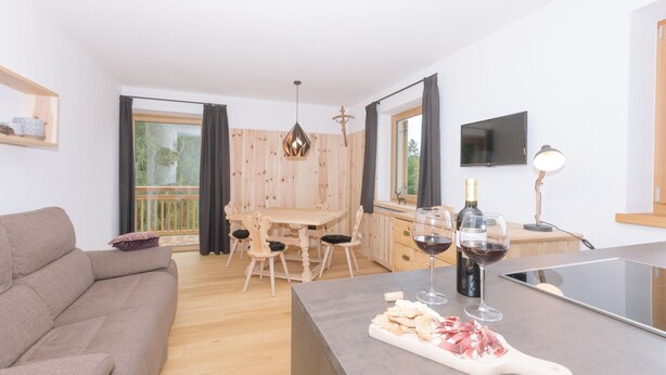 A modern living room with a sofa, dining table, and chairs, a kitchen area with a cutting board and wine glasses, and a TV mounted on the wall. - Holiday Home “Berg Wieserhof” with Mountain View, Balcony and Shared Garden (Aldein)