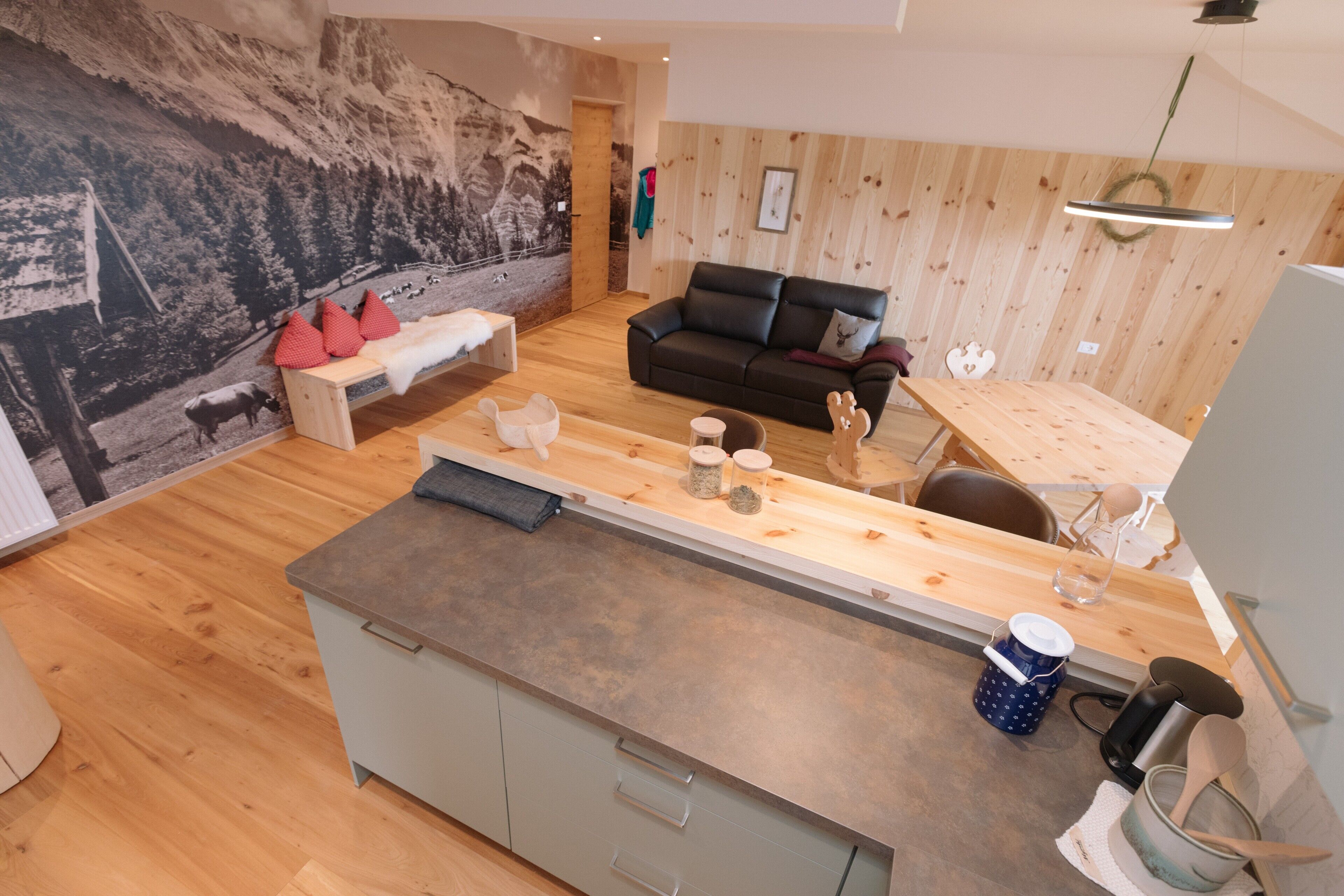 A modern kitchen with a wooden countertop, a dining area with a table and chairs, a black leather sofa, and a mural of a mountain landscape on the wall.