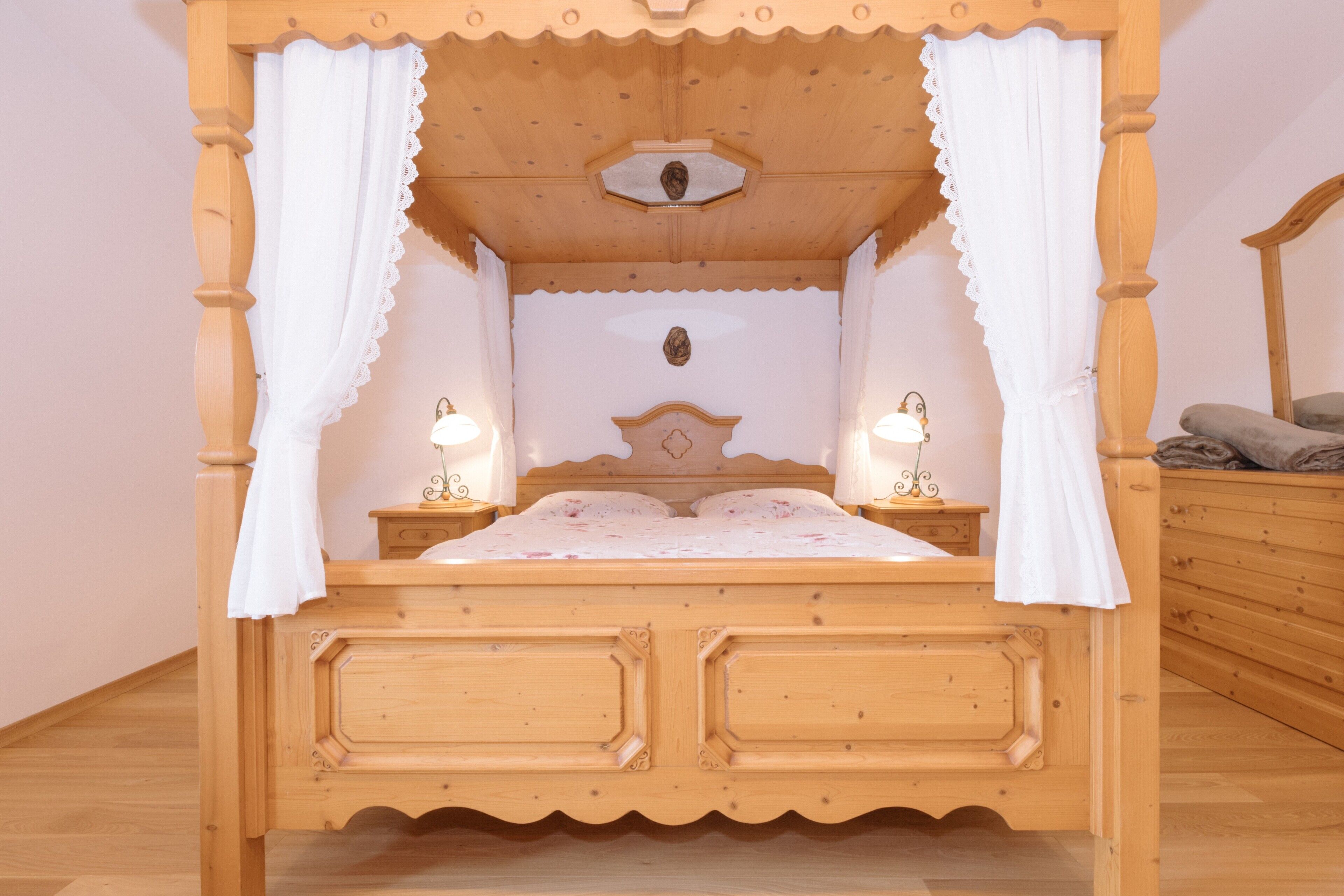 A wooden four-poster bed with white curtains, two bedside tables with lamps, and a wooden dresser.