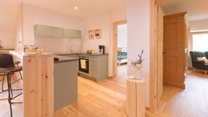 A modern kitchen with a central island, a dining area with a bar stool, and a view into a bedroom with a bed and a window. - Holiday Home “Wies Wieserhof” with Mountain View, Balcony and Shared Garden (Aldein)