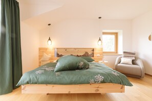 Un dormitorio con cama de madera, ropa de cama verde, dos lámparas de noche, una silla y un ventanal.