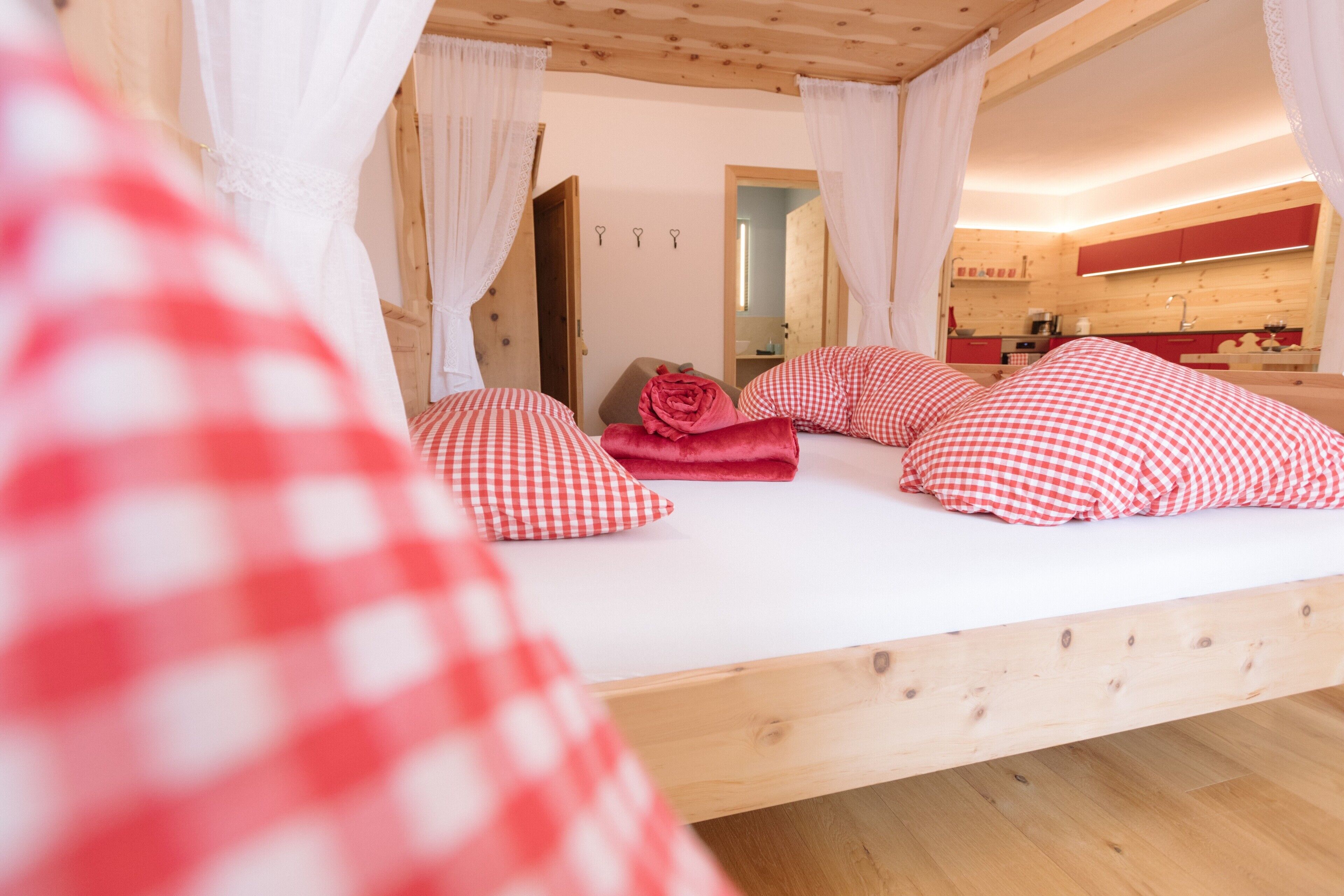 Een houten bed met witte beddengoed en rode geruite kussens, een houten plafond en een zichtbare badkamer met spiegel en wastafel.