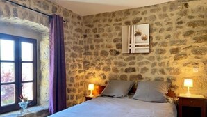 A bedroom with a stone wall, a bed with two pillows, a window with curtains, and two bedside lamps. - Charming Cottage Near Puy-en-Velay with Private Terrace and Modern Amenities (Blavozy)