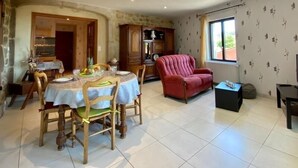 A dining area with a round table set for four, surrounded by wooden chairs. A red armchair is positioned near a window with a view of greenery outside. - Charming Cottage Near Puy-en-Velay with Private Terrace and Modern Amenities (Blavozy)