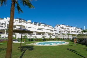 A pool area with a palm tree, umbrellas, and a building with balconies in the background. - Lotus Golf & Beach 2306 (Casares)