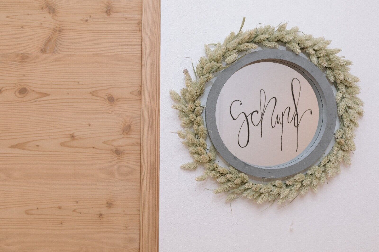 Een houten deur en een rond bord aan de muur, waarop 'schön' staat, omringd door een krans.