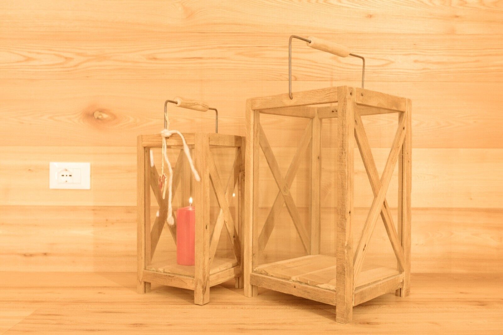 Two wooden lanterns with handles, one with a red candle, against a wooden wall background.