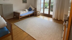 Una habitación con una cama, una mesita pequeña, un radiador y un ventanal grande con cortinas.