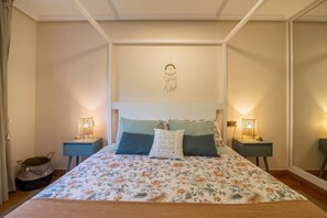 A bedroom with a floral bedspread, two bedside lamps, a dreamcatcher on the wall, and a basket on the floor.