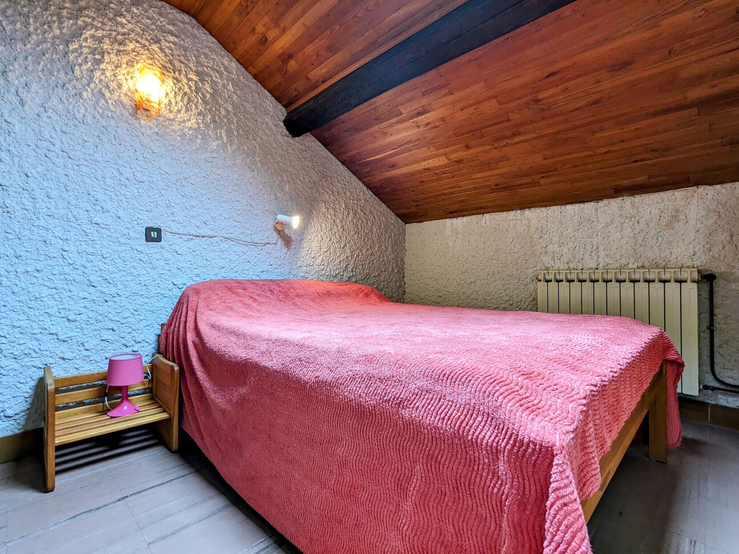 A cozy attic bedroom with a sloped ceiling, a wooden bench, a pink bedspread, and a wall-mounted light fixture.