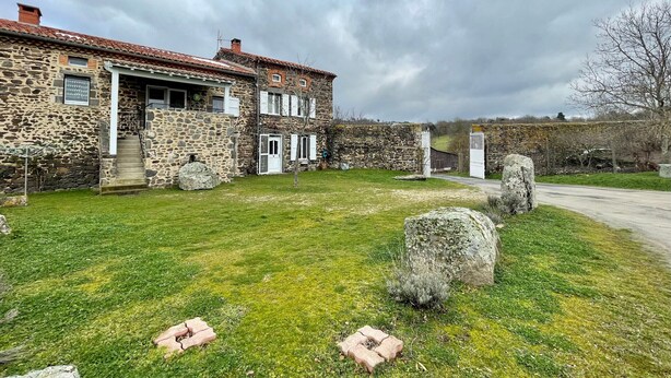Une maison en pierre avec un toit de tuiles, un mur en pierre et une cour verdoyante parsemée de gros rochers.
