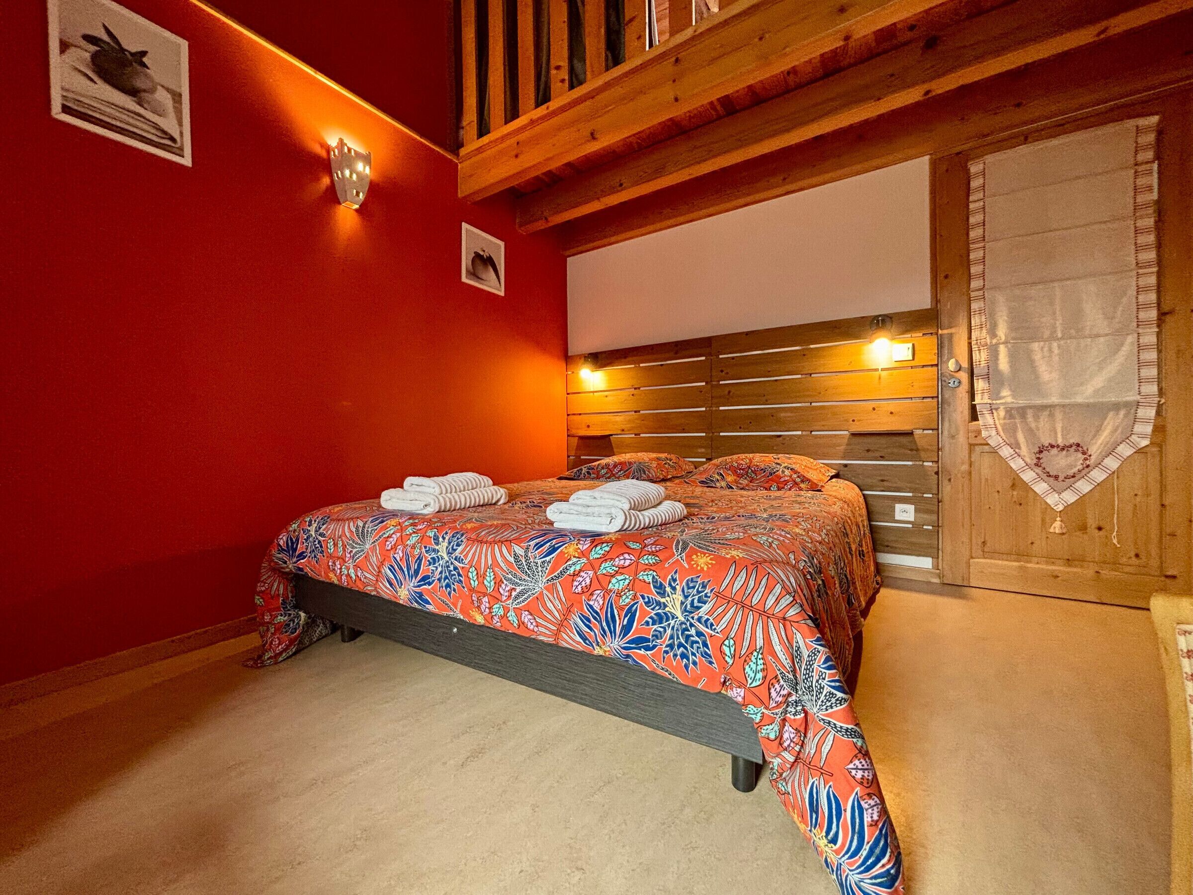 Une chambre à coucher avec une tête de lit en bois, un mur rouge et un lit recouvert d’une courtepointe à motifs floraux et agrémenté de serviettes.