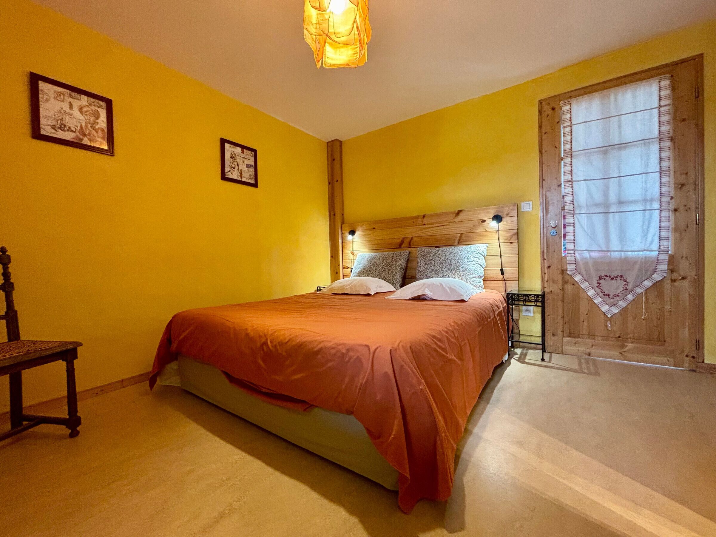 A bedroom with a wooden headboard, a bed with an orange bedspread, two framed pictures on the wall, and a window with a heart-shaped curtain.