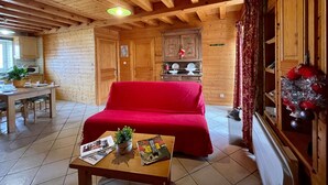 L’intérieur d’un chalet confortable, avec un canapé rouge, une table à manger entourée de chaises, un coin cuisine et une fenêtre agrémentée de rideaux.