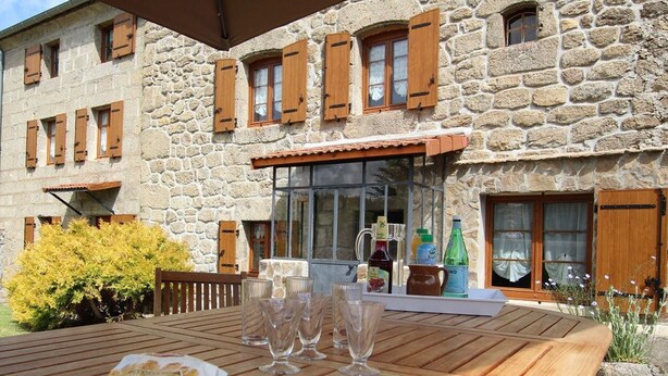 A stone building with wooden shutters, a patio table with food and glasses, and a bottle of wine.