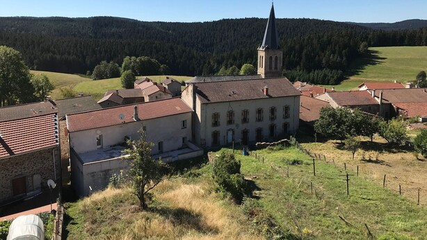 A rural village with a church featuring a tall spire, surrounded by houses with red-tiled roofs and a hilly forested backdrop.