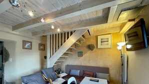 Una habitació acollidora amb sostre de fusta, una escala, un sofà, una taula i una televisió.