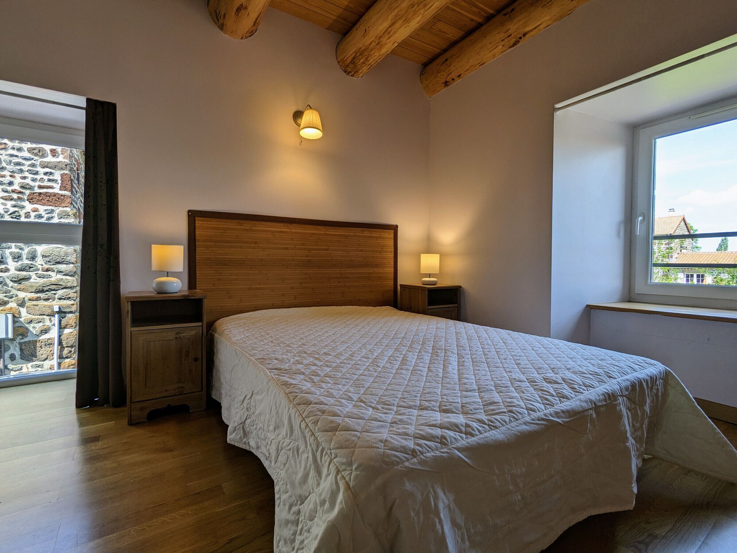 Een slaapkamer met een groot bed, houten hoofdbord, twee bedlampjes, een stenen muur en een raam met uitzicht.