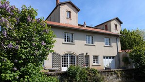 Une maison à deux étages, avec un toit recouvert de tuiles rouges, un mur en pierre et un jardin où poussent des fleurs violettes.