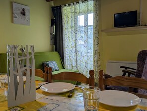 Un coin repas avec une table dressée pour deux personnes, un canapé vert, un téléviseur et une fenêtre ornée de rideaux à motifs.