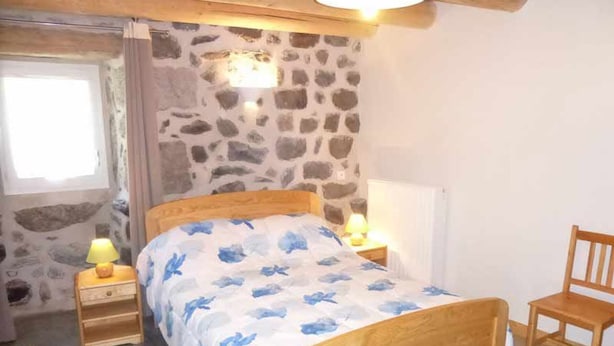 A bedroom with a stone wall, a wooden bed, two bedside lamps, a wooden chair, and a window with curtains. - Charming Village Farmhouse Sleeps 8 – Near Allier Gorges & Pradelles (Rauret)