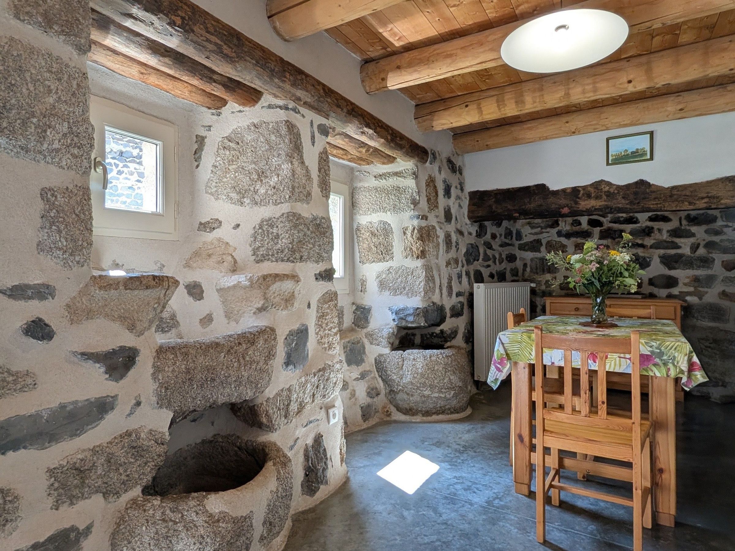 Un intérieur rustique avec des murs en pierre, une table et des chaises en bois, un mur en pierre percé d’une fenêtre et un radiateur.