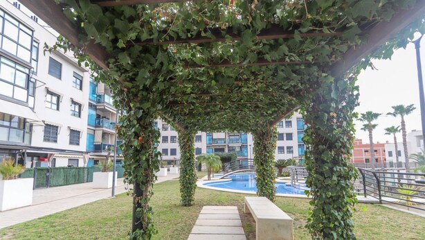 Un espacio exterior techado con un sendero que lleva a una piscina, rodeado de vegetación y edificios modernos.