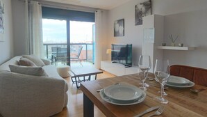Una sala de estar moderna con un sofá, una mesa de comedor dispuesta con platos y vasos, un televisor y vistas a la ciudad a través de grandes ventanales.