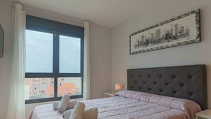 Un dormitorio con un ventanal grande, una cama con cabecera capitonada y un cuadro de paisaje urbano en la pared.