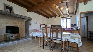 Zona pranzo con tavolo e sedie in legno, camino in pietra, televisore su un supporto e cucina sullo sfondo.