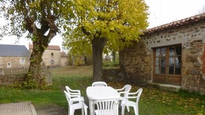 Un patio con tavolo e sedie bianche, circondato da muri in pietra e un albero con foglie gialle.