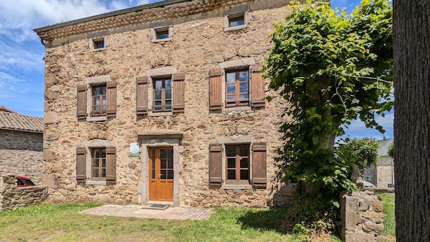 Un edificio in pietra con una porta e delle persiane di legno, circondato da erba e un albero.