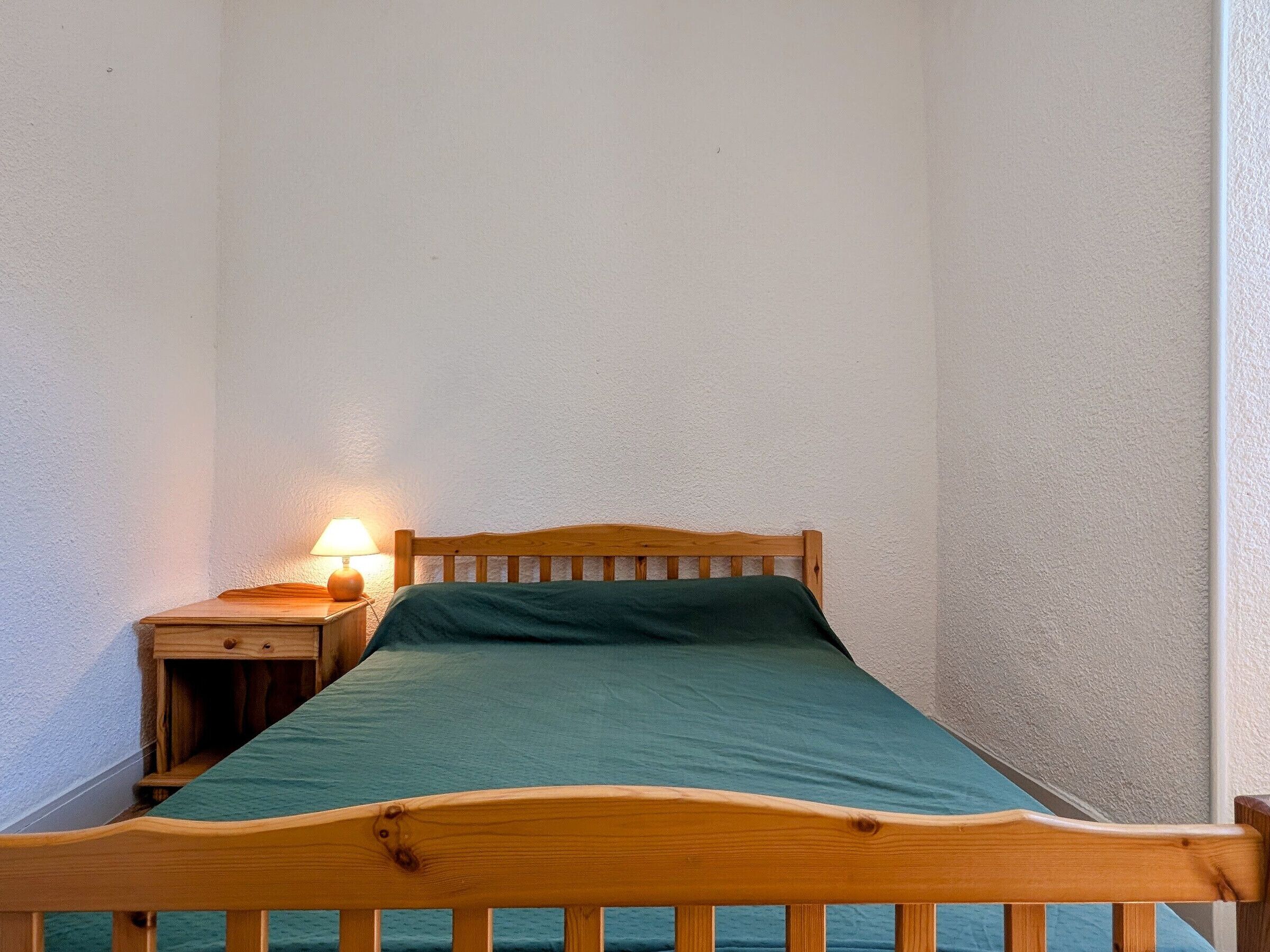 A single bed with a wooden frame and a green bedspread in a room with white walls and a small bedside table with a lamp.