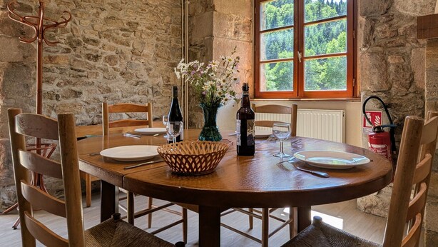 Un comedor de paredes de piedra con una mesa redonda, sillas, botellas de vino, un jarrón con flores y una ventana que da a un bosque.